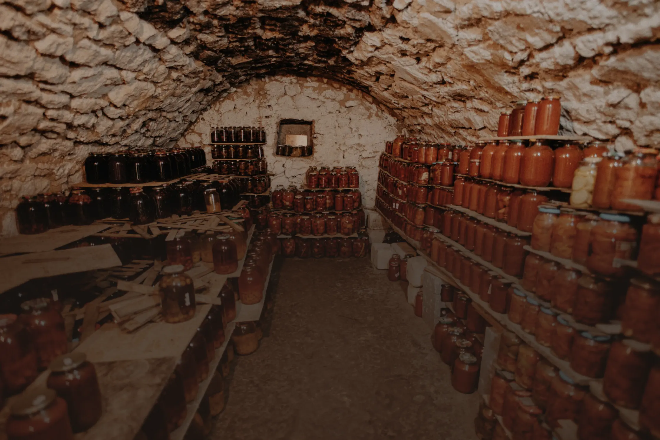 Une cave voûtée en pierre sèche dont les planches portent des rangées de bocaux de conserves.