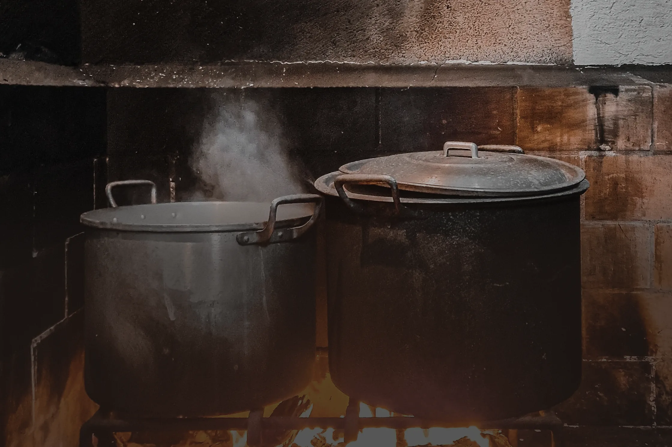 Deux grandes marmites qui mijotent sur un feu de bois, dans le foyer maçonné d’une cuisine de ferme, sous la porte de fonte du four à pain.