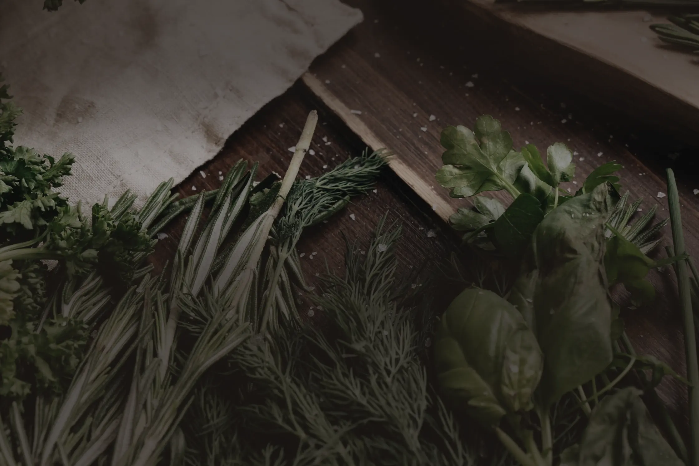 Des herbes fraîches coupées — romarin, aneth, persil, basilic — étalées sur une table de bois sombre à côté d’un bol de poivre.
