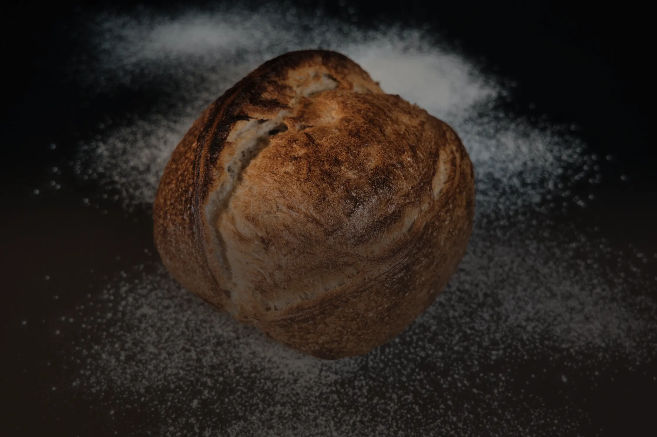 Une miche de pain au levain à la croûte fendue, posée sur un plan sombre saupoudré de farine.