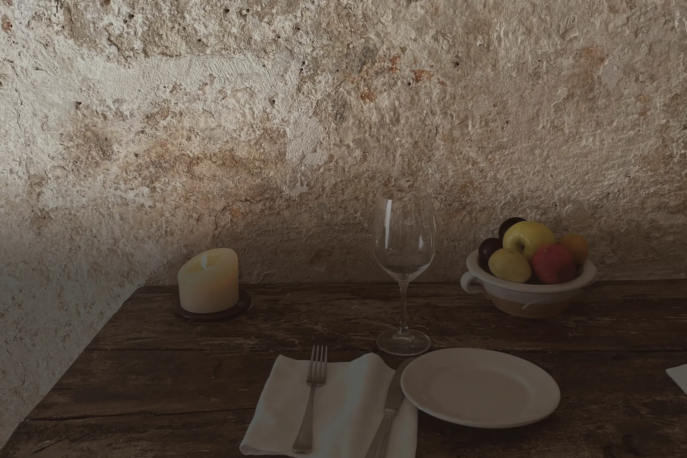 Un couvert dressé sur une table de bois brut devant un mur de chaux écaillé : une bougie, un verre, une coupe de fruits.